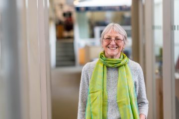 Angela Towle stands facing the camera and smiling wearing a bright green scarf over a grey sweater in the hallway at the UBC Learning Exchange.